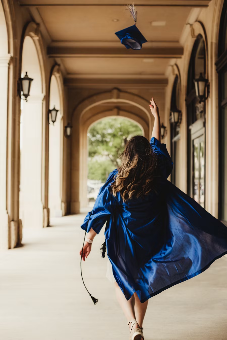 Person in a blue graduation gown throwing a blue graduation cap in an archway.